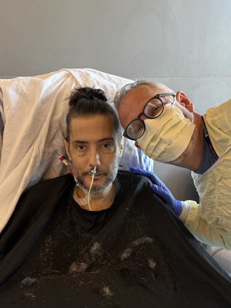 Nathaniel and Richard. Nathaniel is still in the recliner covered with a white sheet. His cape is covered with hair. His shorter hair is now in a man bun. His beard is much shorter. Richard is wearing a yellow mask and gown as well as dark-rimmed gasses.